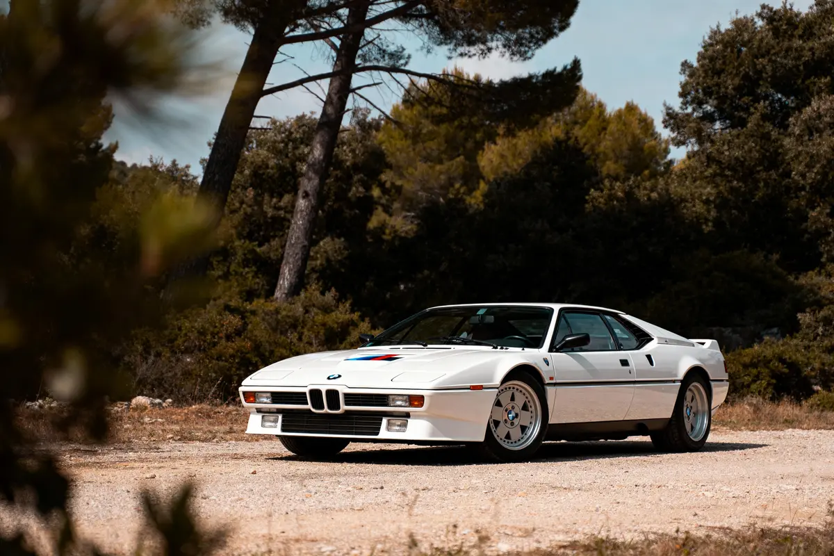Voiture sportive blanche stationnée sur chemin de terre, entourée d'arbres et de végétation luxuriante.