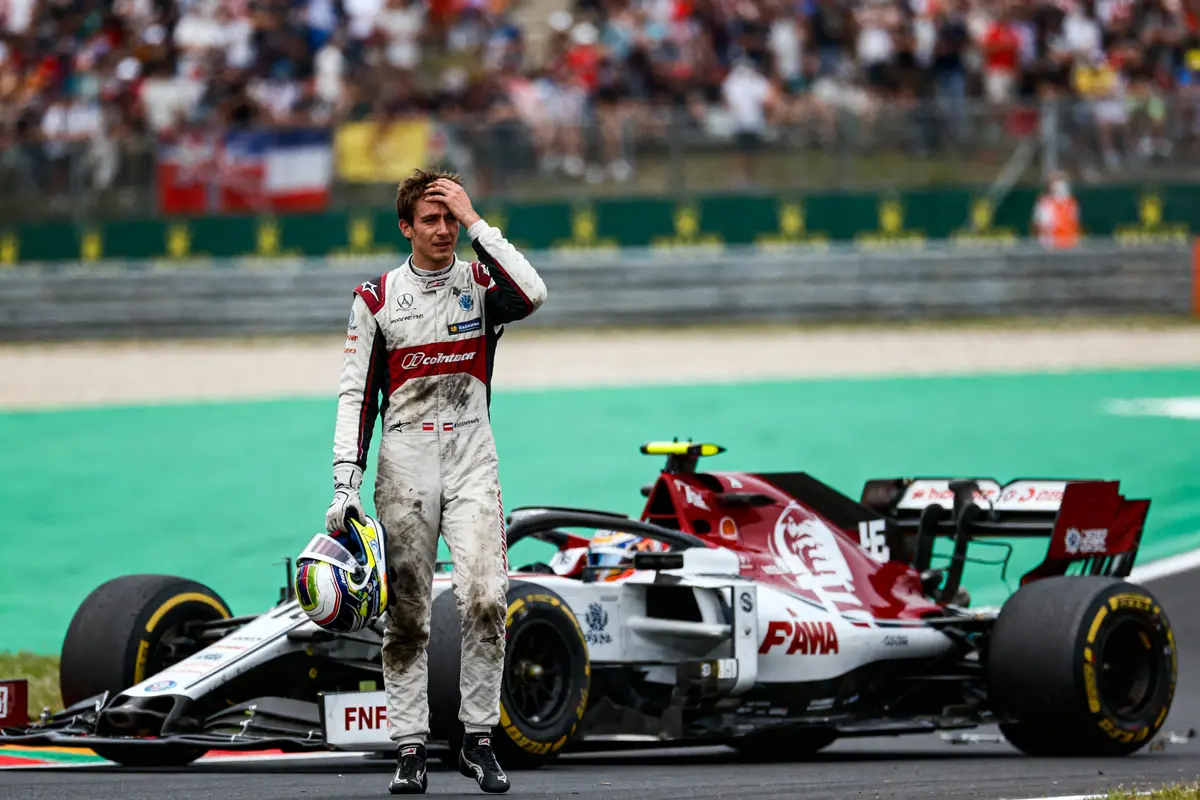 Pilote de course en combinaison blanche, tenant son casque, marche devant une voiture de Formule 1 endommagée.