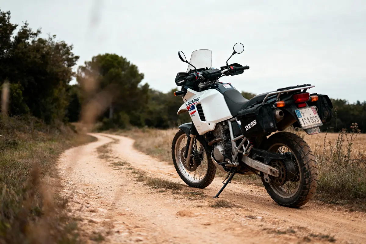 Moto tout-terrain blanche sur un chemin de terre sinueux, entourée de végétation et ciel nuageux.
