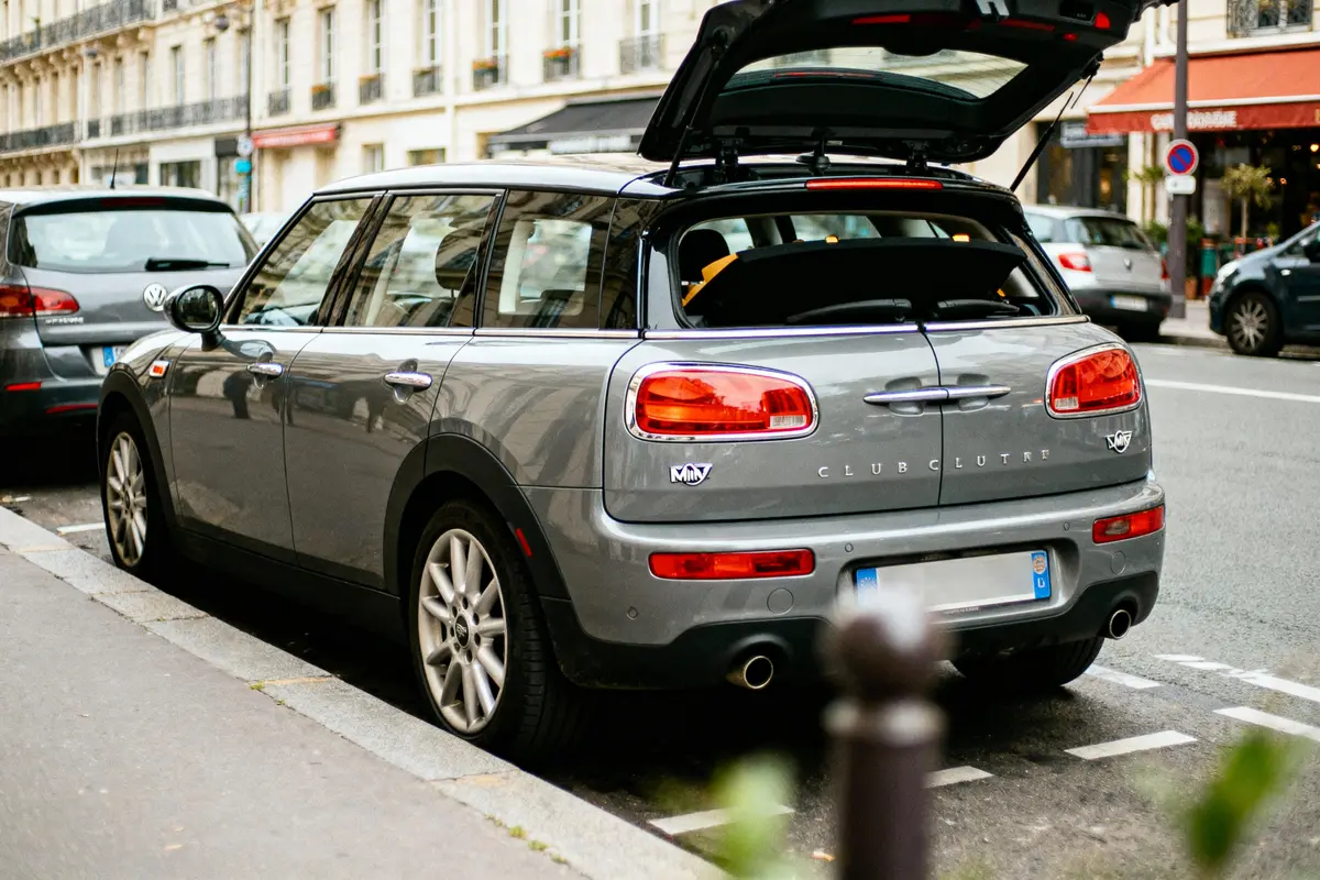 Voiture grise Mini Clubman stationnée, coffre ouvert, rue urbaine avec bâtiments en arrière-plan.
