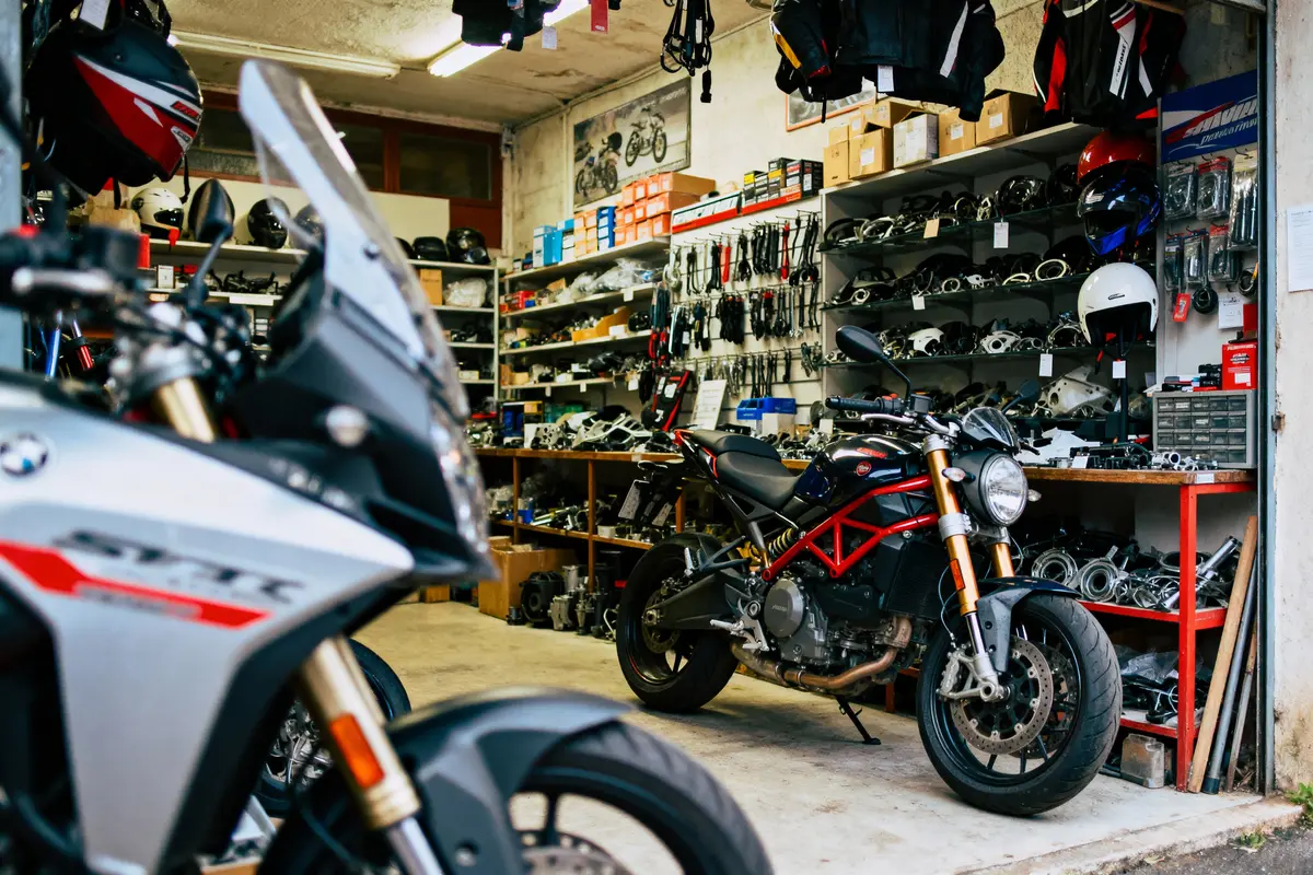 Atelier de motos avec divers modèles exposés, outils et accessoires en arrière-plan sur des étagères. Éclairage naturel.