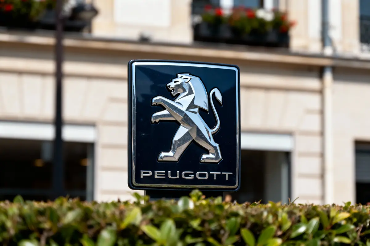 Logo de voiture avec lion métallique sur fond de bâtiment parisien, texte "PEUGEOTT" en dessous, partiellement caché par des buissons.