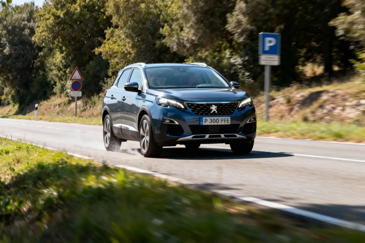Un SUV Peugeot bleu roule sur une route, entouré d'arbres, avec des panneaux de circulation visibles à l'arrière-plan.