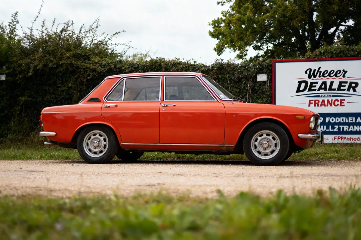 Voiture classique orange stationnée devant un panneau publicitaire, chemin de gravier et haies en arrière-plan.