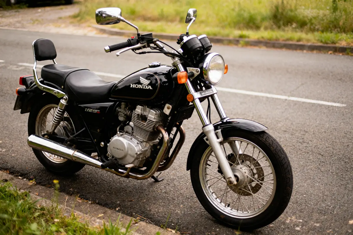 Moto noire Honda CB125T stationnée sur une route, gazon en arrière-plan. Vue de côté, lumière naturelle.