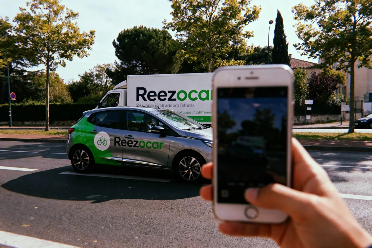 Voiture Reezocar photographiée avec smartphone en ville, camion en arrière-plan, arbres entourant la scène.