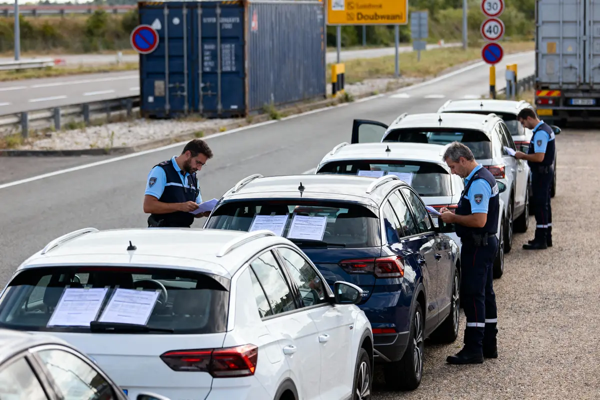 Agents vérifient des documents sur voitures alignées à un contrôle routier, zone industrielle en arrière-plan.