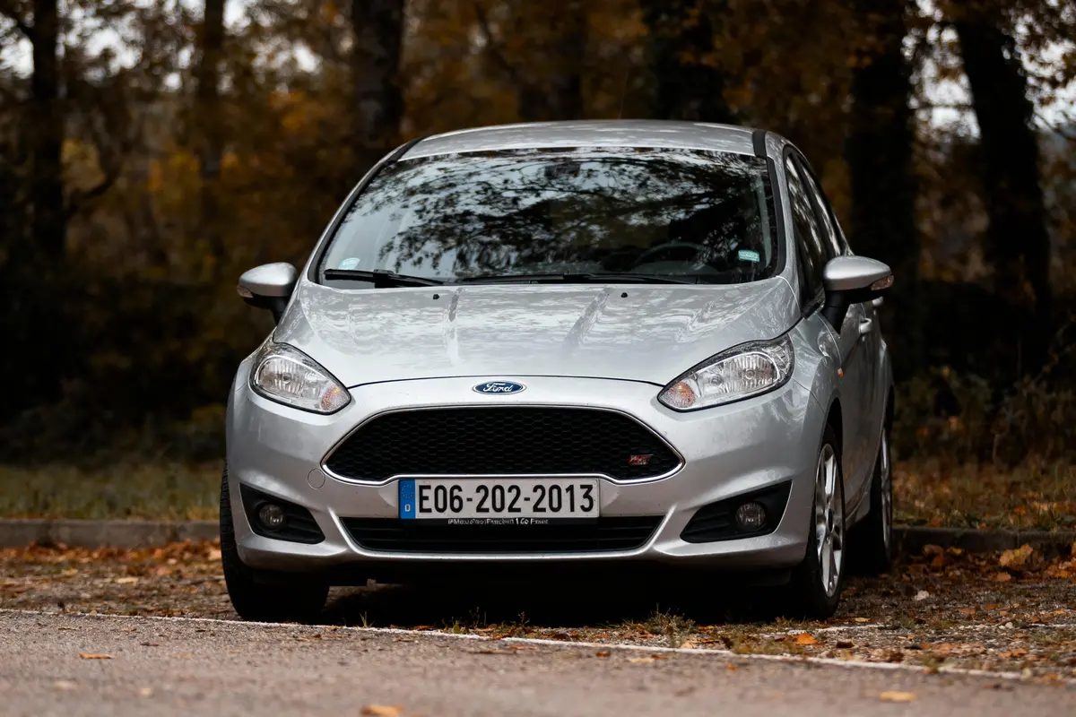 Voiture Ford argentée garée sur route, fond forestier automnal avec feuilles marron et arbres. Plaque visible.