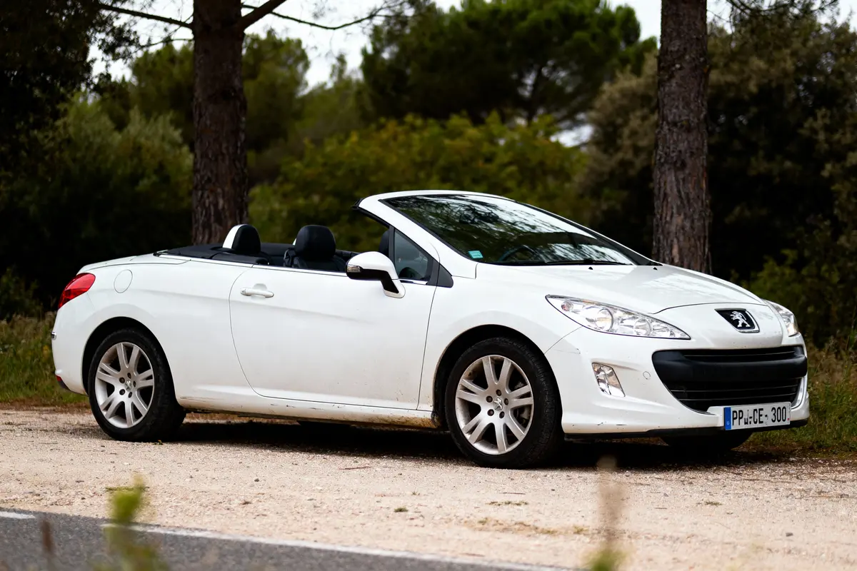 Voiture décapotable blanche Peugeot stationnée sur chemin de terre, fond de végétation et arbres.