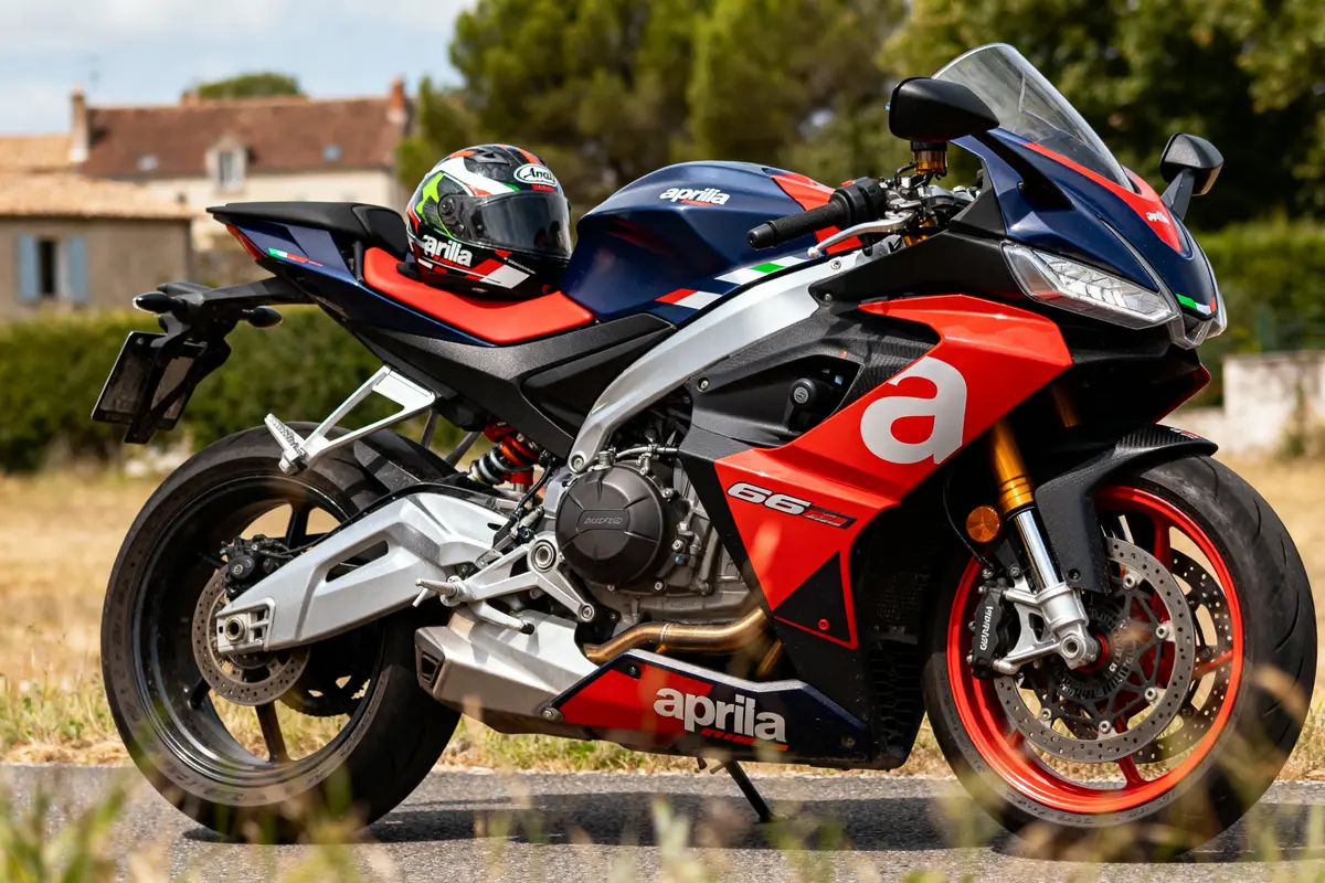 Moto Aprilia sportive rouge, noire, blanche, stationnée sur route, casque assorti posé dessus, maisons et arbres en arrière-plan.