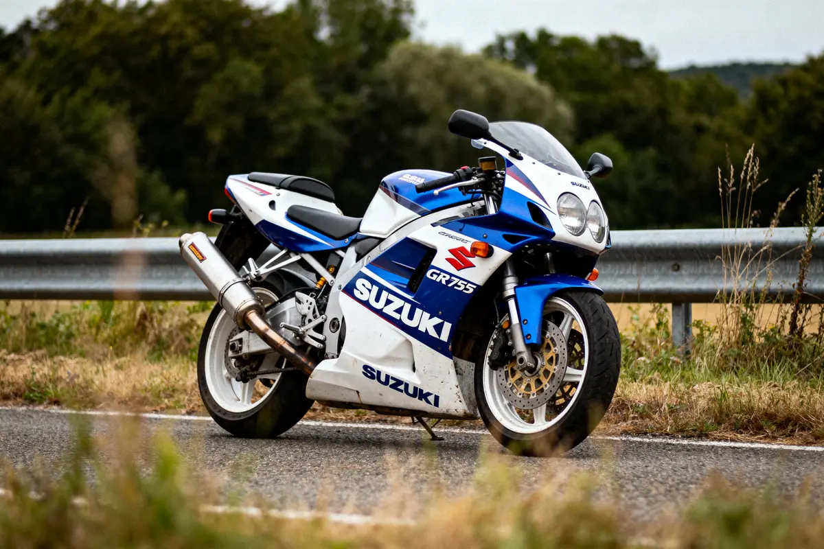 Moto Suzuki bleue et blanche stationnée sur route, avec végétation floue en arrière-plan, garde-corps métallique.