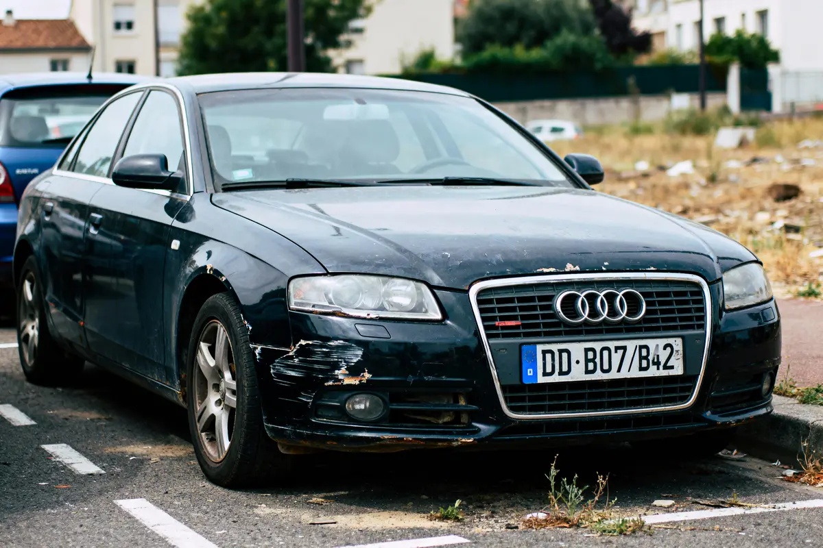 Voiture noire Audi endommagée, garée en extérieur, peinture abîmée et rayures visibles sur le pare-chocs avant.