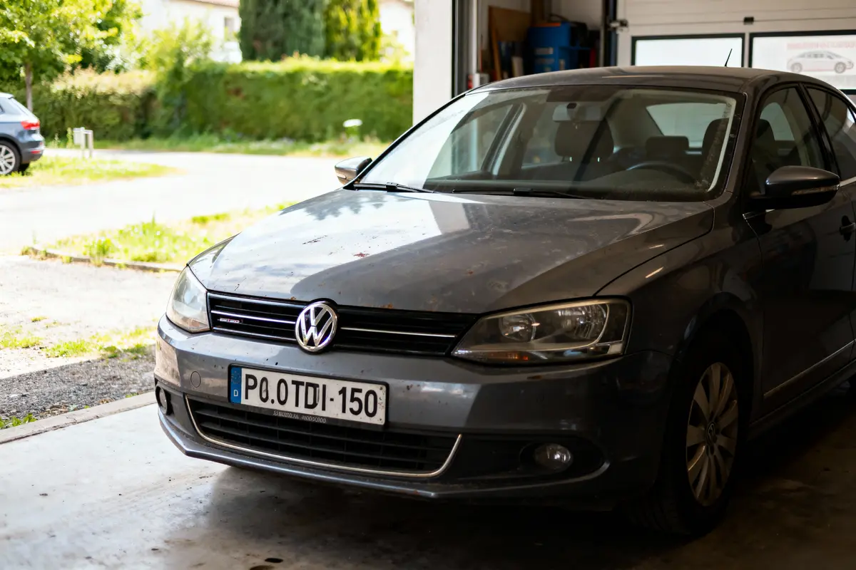 Voiture Volkswagen grise stationnée dans un garage, partiellement ensoleillé, avec végétation en arrière-plan.