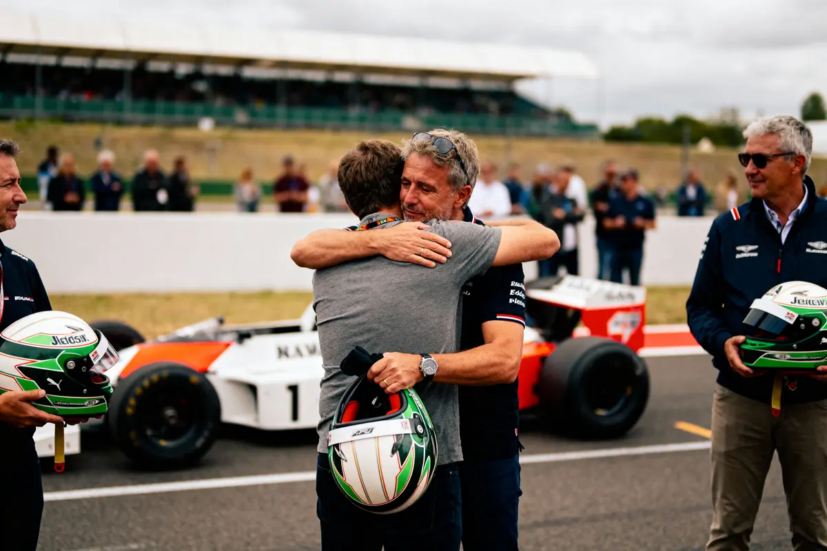 Deux hommes s'embrassent devant une voiture de course, casques à la main, sur un circuit automobile.