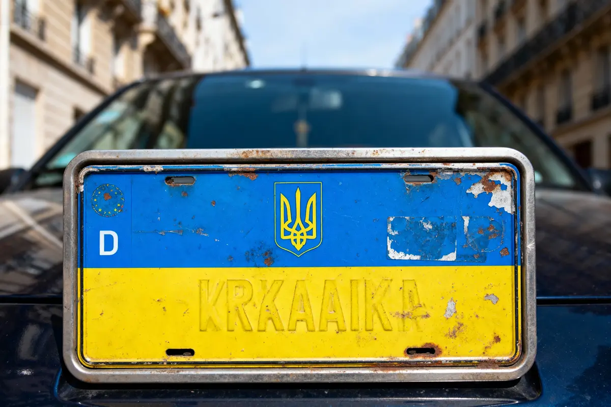 Plaque d'immatriculation usée, aux couleurs ukrainiennes, sur fond de voiture noire dans une rue urbaine.