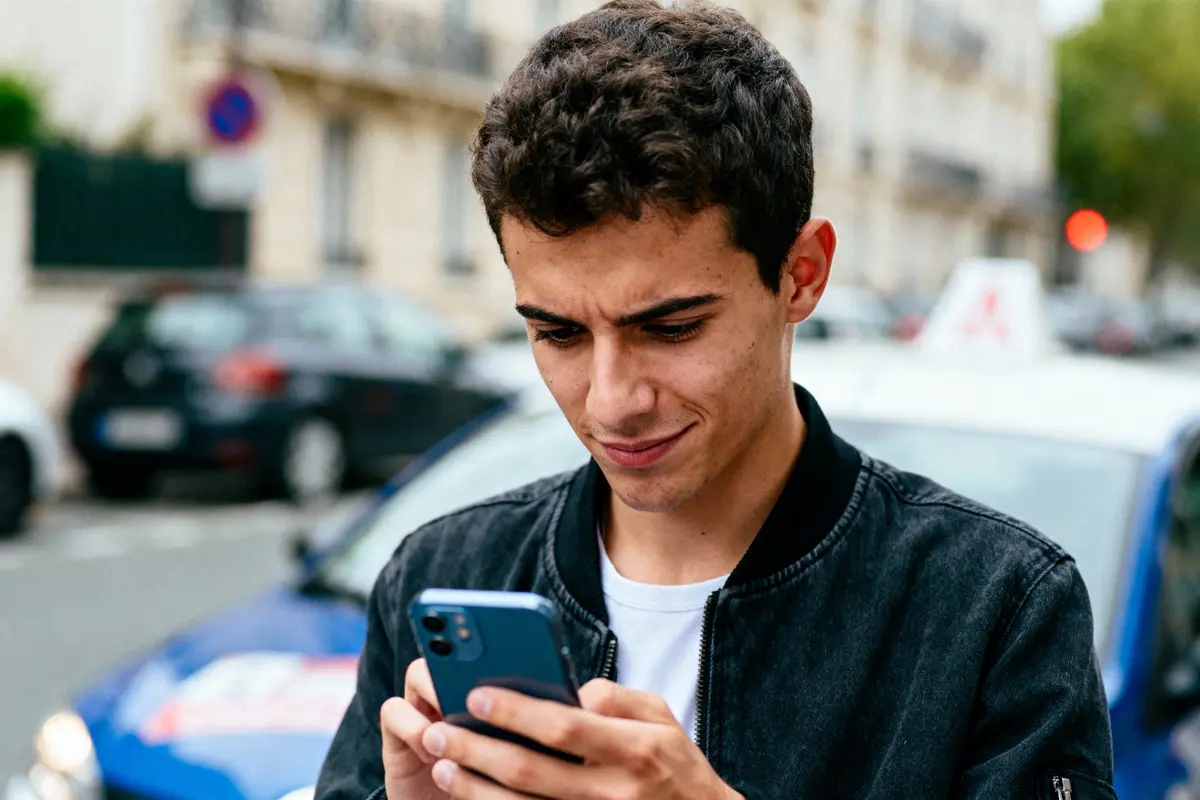 Jeune homme concentré utilise son smartphone dans une rue urbaine, voitures en arrière-plan flou.
