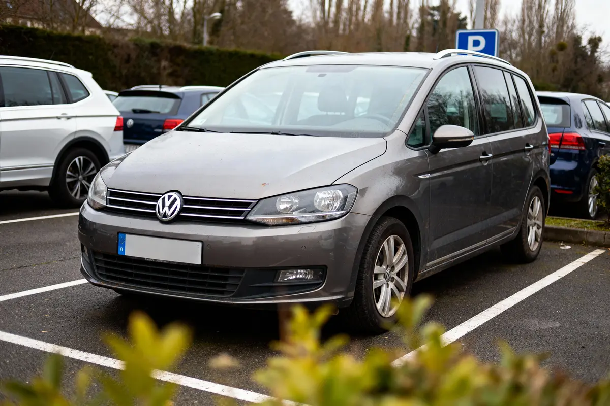 Voiture Volkswagen grise garée sur un parking, entourée d'autres véhicules et de végétation.