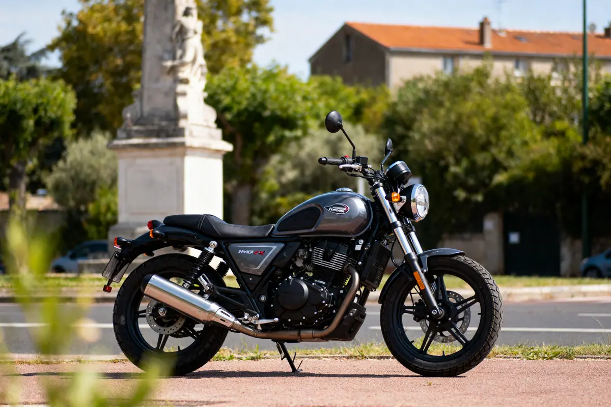 Moto de style rétro Hyosung GV125S garée devant un monument en arrière-plan urbain verdoyant.