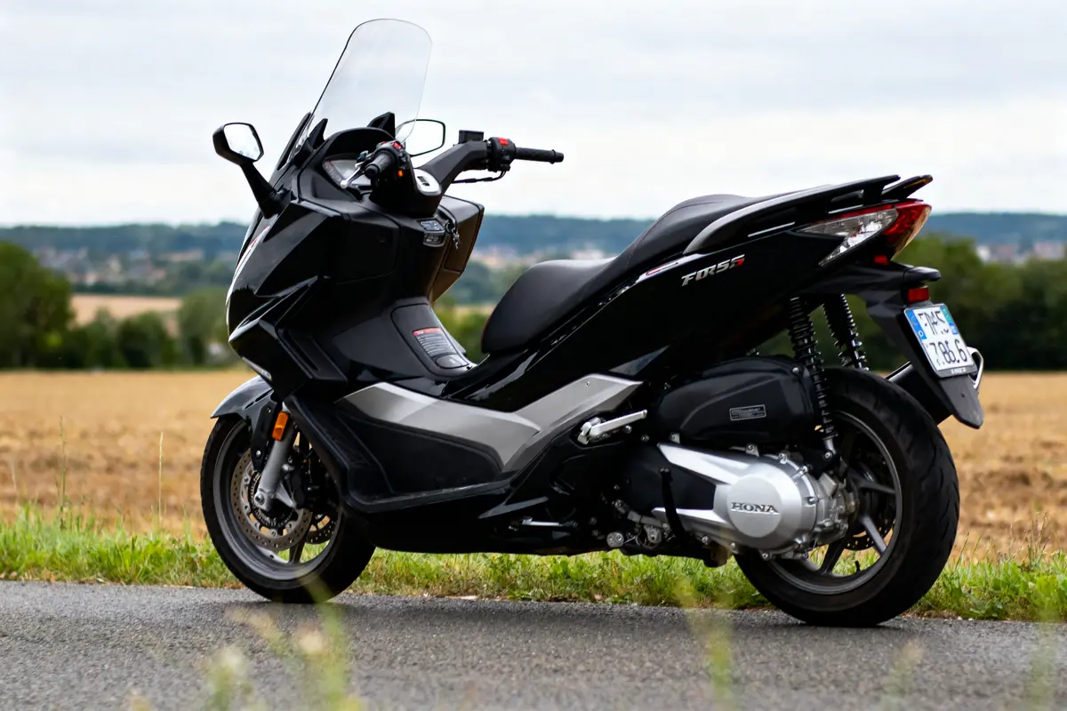 Scooter noir Honda Forza garé sur route de campagne avec paysage campagnard en arrière-plan.