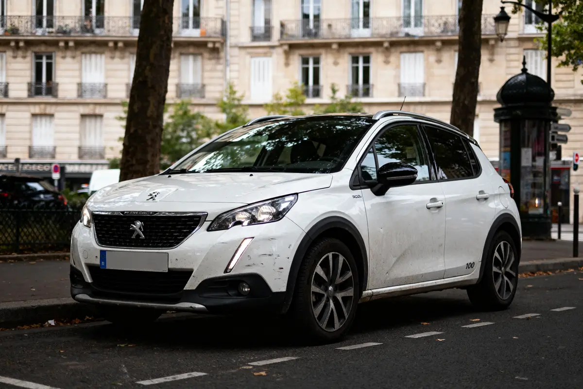 SUV Peugeot blanc garé en ville devant immeuble, arbres et trottoir. Journée nuageuse.