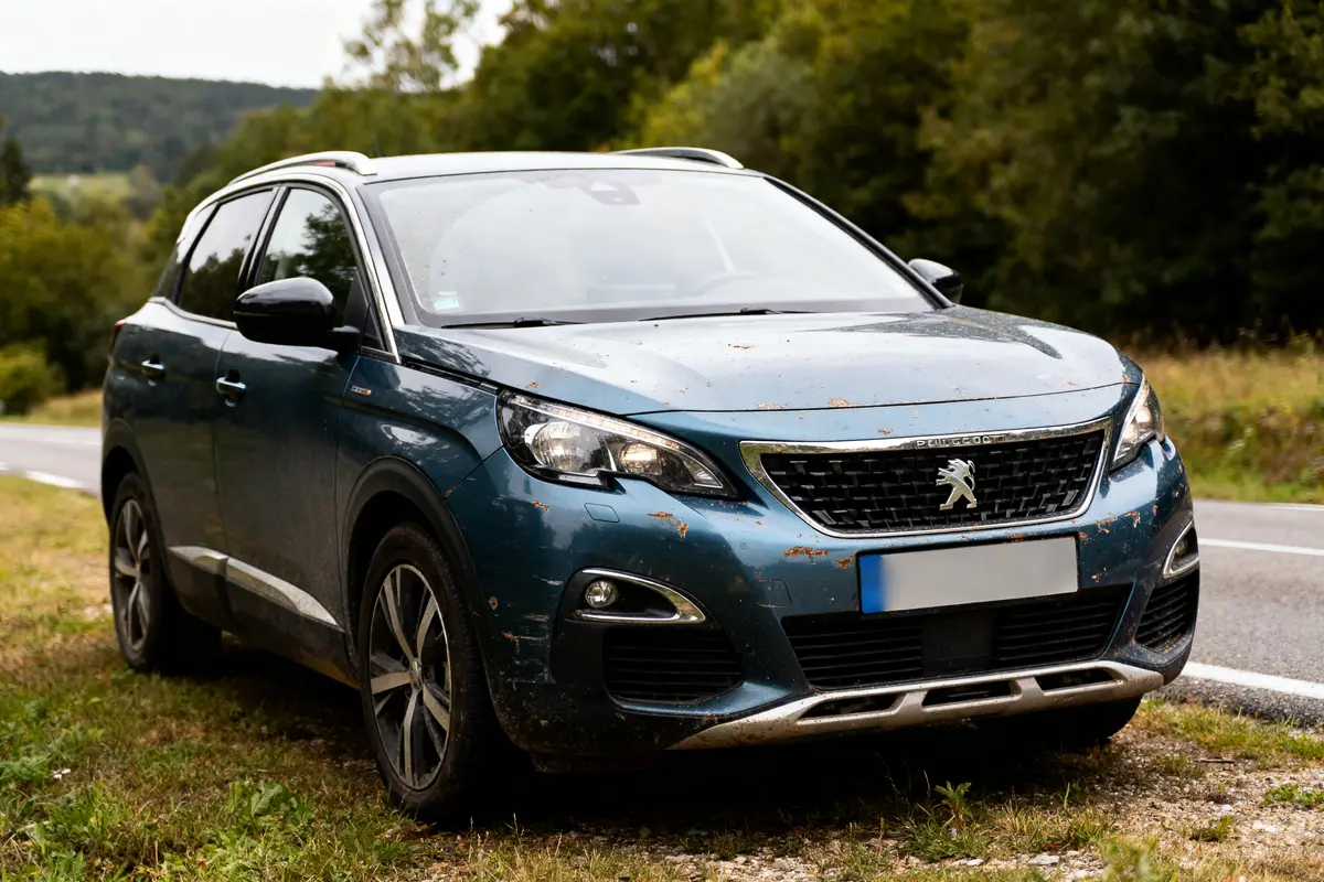 SUV bleu Peugeot garé sur bas-côté herbeux, route et paysage boisé en arrière-plan.
