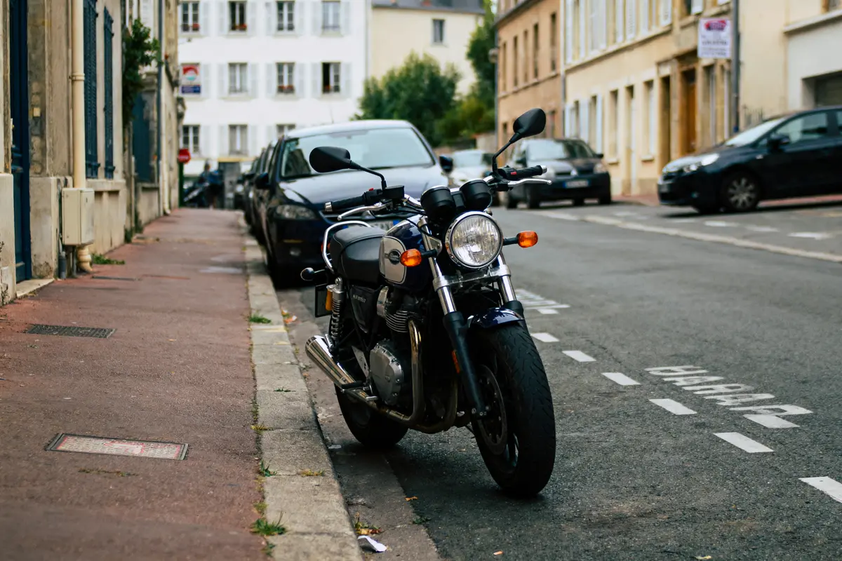 Moto stationnée sur trottoir urbain, rue calme bordée de voitures et bâtiments résidentiels anciens.