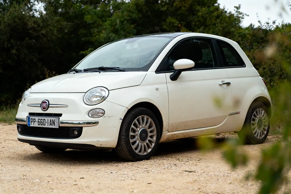 Voiture Fiat 500 blanche, stationnée sur chemin terreux, entourée de végétation verdoyante en arrière-plan.
