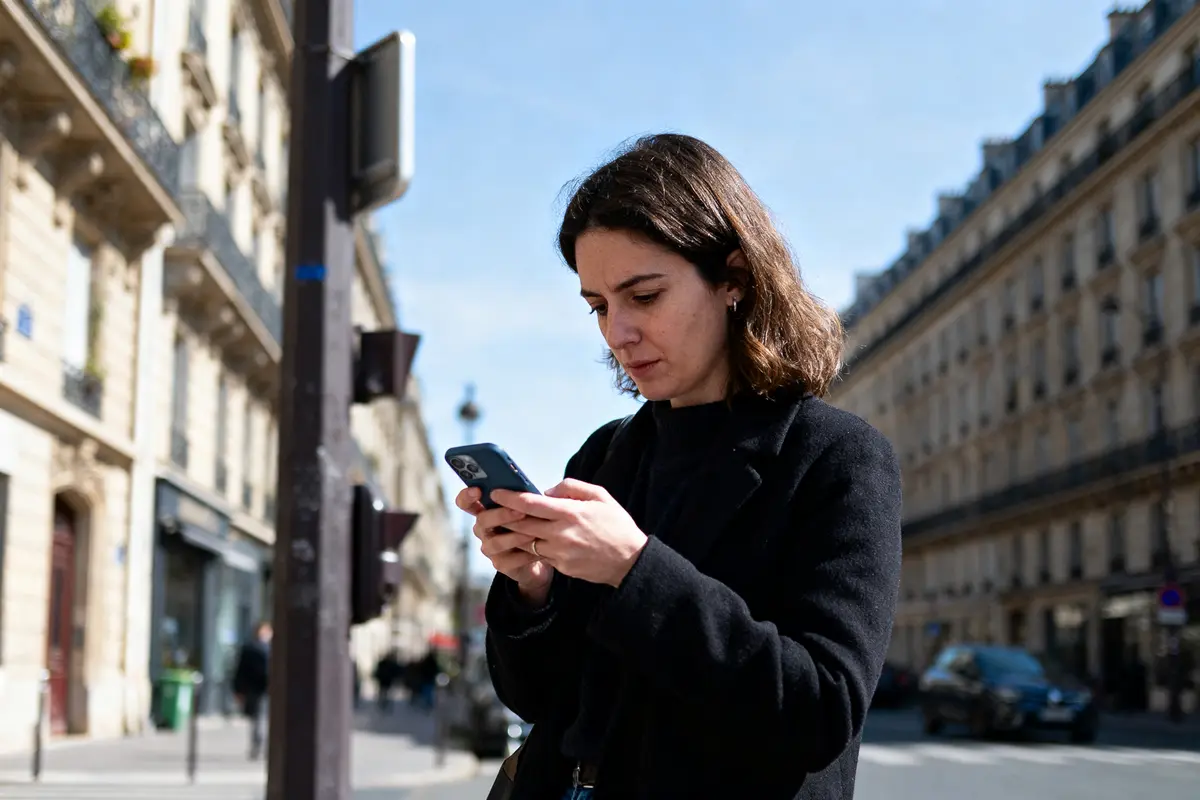 Femme regardant son téléphone dans une rue urbaine avec bâtiments en arrière-plan, ciel bleu.