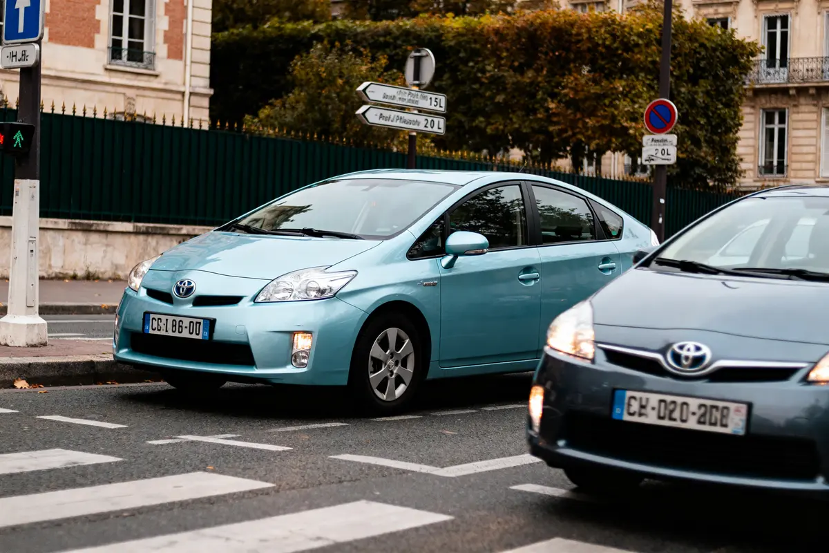 Deux voitures Toyota Prius hybrides sur la route, devant un passage piéton, avec panneaux directionnels et bâtiments en arrière-plan.