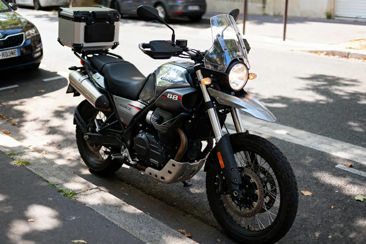Moto de type adventure stationnée sur une rue, équipée d'un top case et de protections avant.