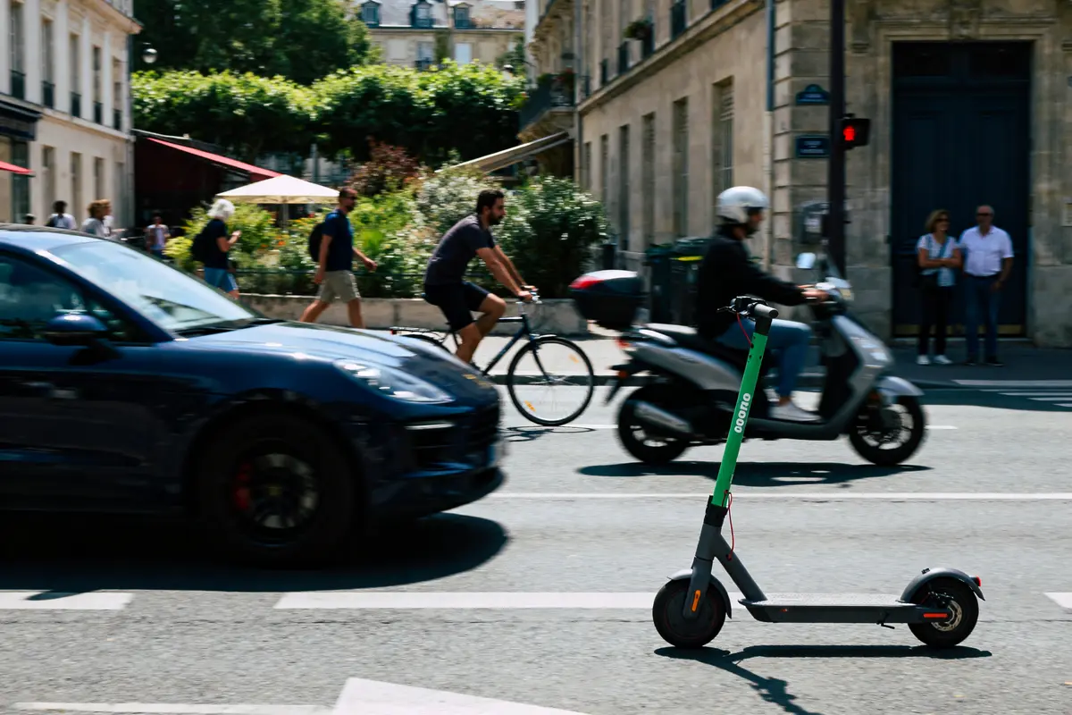 Trottinette électrique, voiture, cycliste et scooter sur une rue urbaine animée en journée ensoleillée.