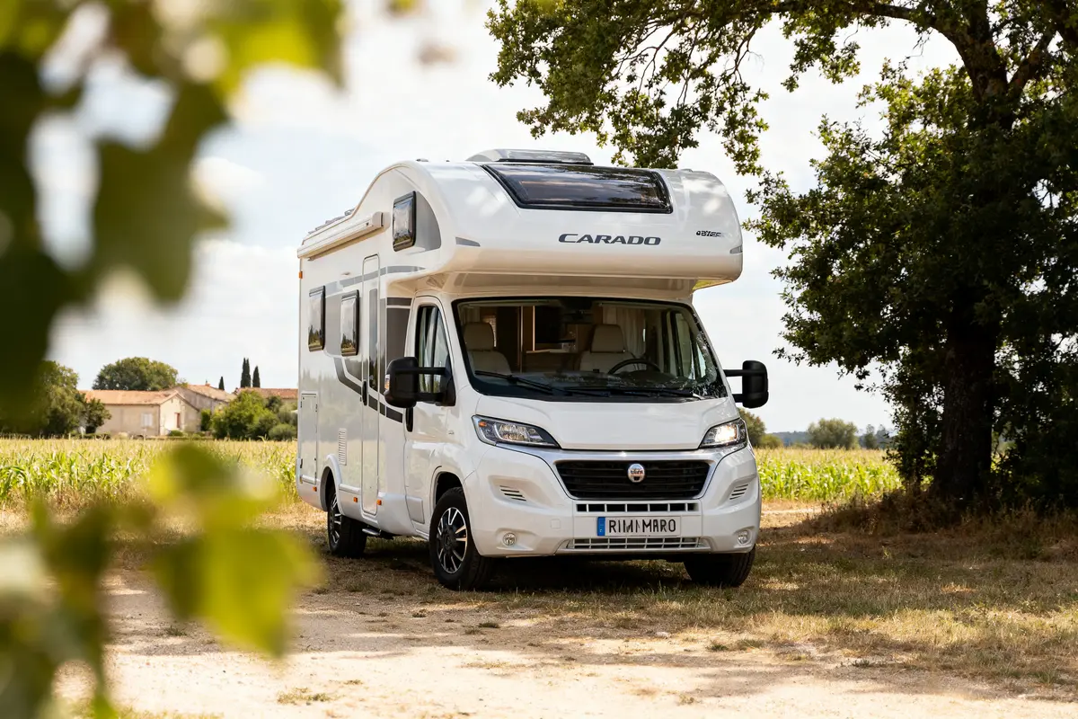 Camping-car blanc Carado stationné dans un champ ensoleillé avec des arbres et une maison en arrière-plan.