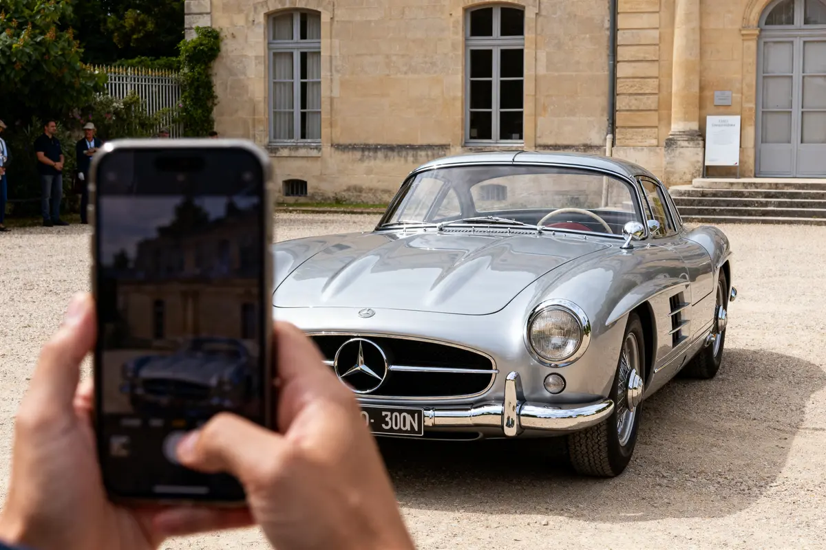 Une voiture de sport classique argentée photograhiée devant un bâtiment historique avec smartphone au premier plan.