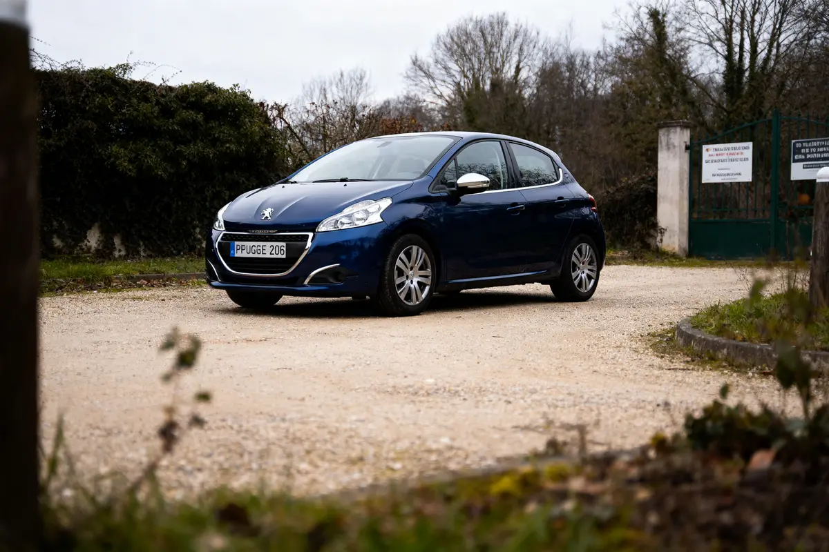 Voiture bleue Peugeot garée sur chemin de gravier, entourée d'arbres et de buissons en arrière-plan.