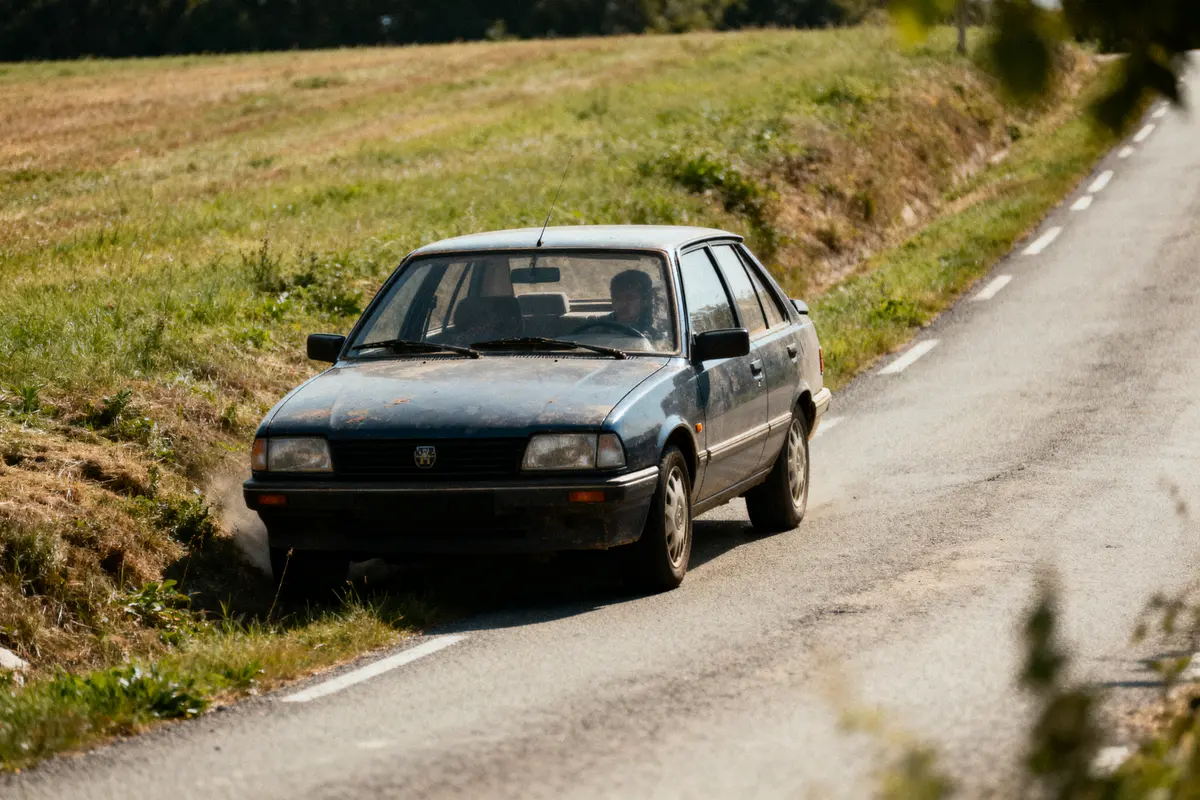 Voiture ancienne bleue roulant sur une route de campagne, entourée de verdure.