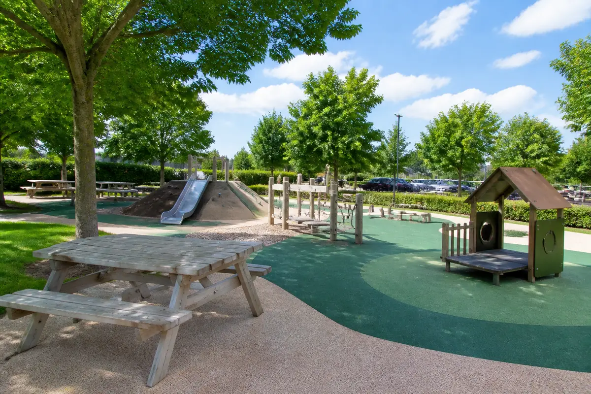 Aire de jeux extérieure avec toboggan, balançoires et tables de pique-nique, entourée d'arbres et de pelouses.