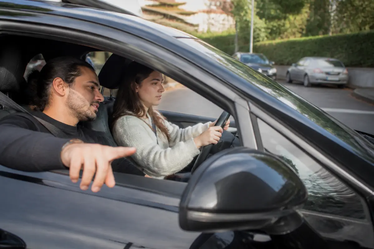Une femme conduit une voiture, accompagnée d'un instructeur qui lui donne des conseils.